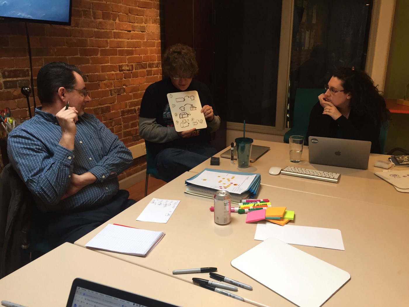 3 people sit at a table, one of whom holds up a drawing.