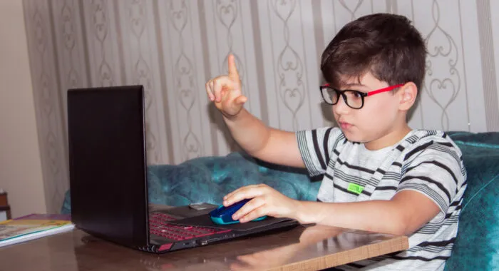 Young boy with glasses and striped shirt at a laptop with right hand raised and pointer finger up