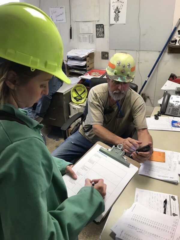 Woman with a hardhat and clipboard watching a man with a hardhat use a mobile device
