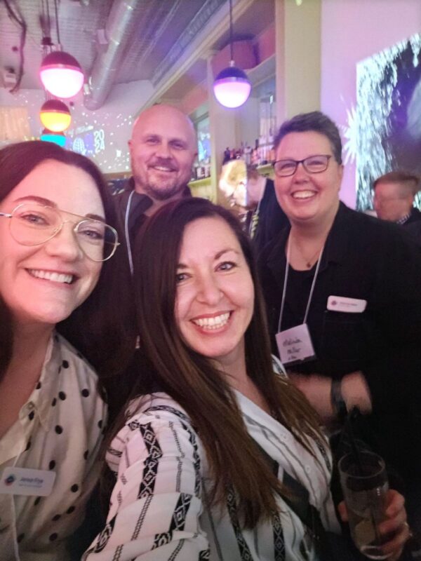 four people smiling at the camera in a selfie, inside a colorful bar at a networking event