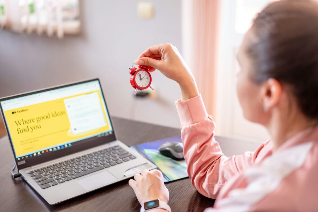 A woman sitting at her laptop holding a small red alarm clock