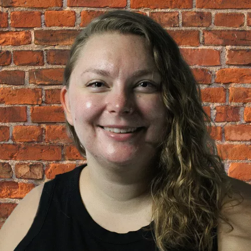 Natalie Semczuk, a woman with brown hair, wearing a black shirt, standing against a brick wall and smiling 
