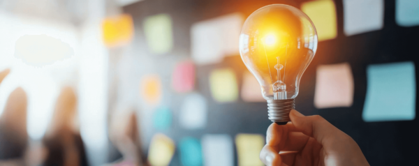 a hand holds up an illuminated lightbulb against a blurred backdrop of a wall covered in sticky notes