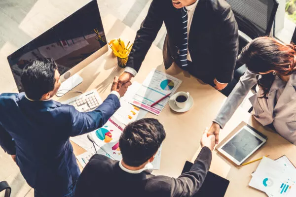 Overhead view of four business professionals in formal attire shaking hands around a desk covered with charts, graphs, and digital devices, symbolizing a successful merger or business agreement.
