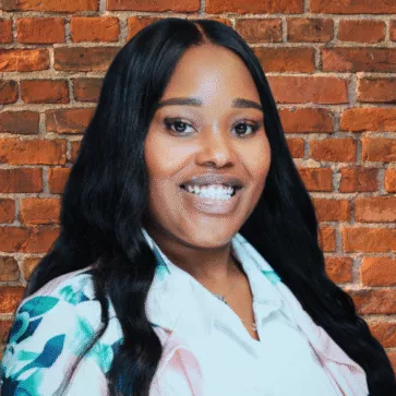 Professional headshot of Lottiah Smith smiling in front of a brick wall background, wearing a white blouse with a patterned blazer.