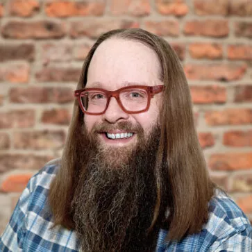 Joe Tower, A smiling man with long straight brown hair, a full beard, and red glasses, wearing a blue plaid shirt, standing in front of a brick wall.