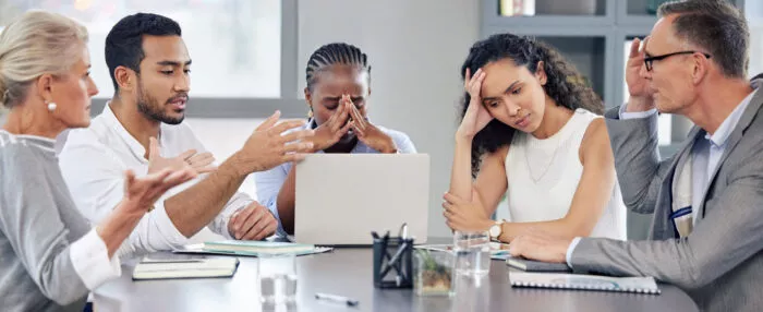 Five people sitting at an office desk with laptops and notebooks appearing to be frustrated and tired.