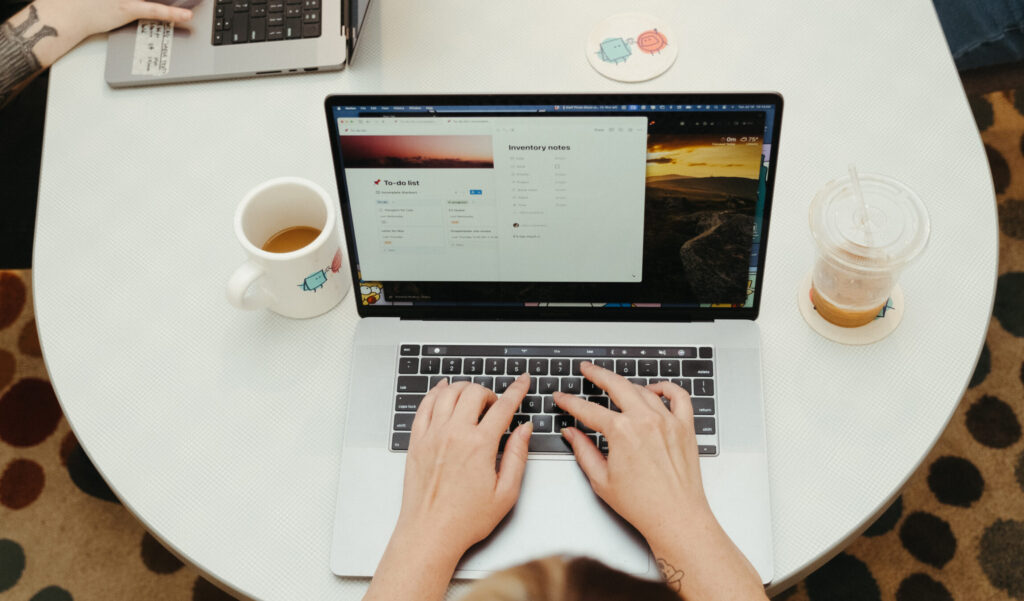 A person typing on a laptop at a table. The table also has a mug of coffee and plastic cup of coffee on it. 