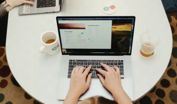 A person typing on a laptop at a table. The table also has a mug of coffee and plastic cup of coffee on it. 