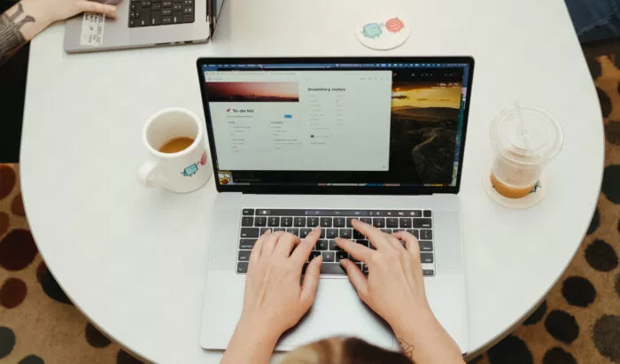 A person typing on a laptop at a table. The table also has a mug of coffee and plastic cup of coffee on it. 