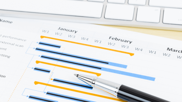 Printed Gantt chart overlaid with a pen, sitting on a desk near a computer keyboard