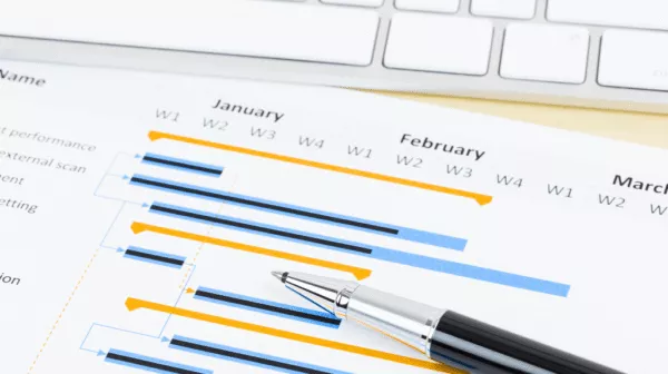 Printed Gantt chart overlaid with a pen, sitting on a desk near a computer keyboard